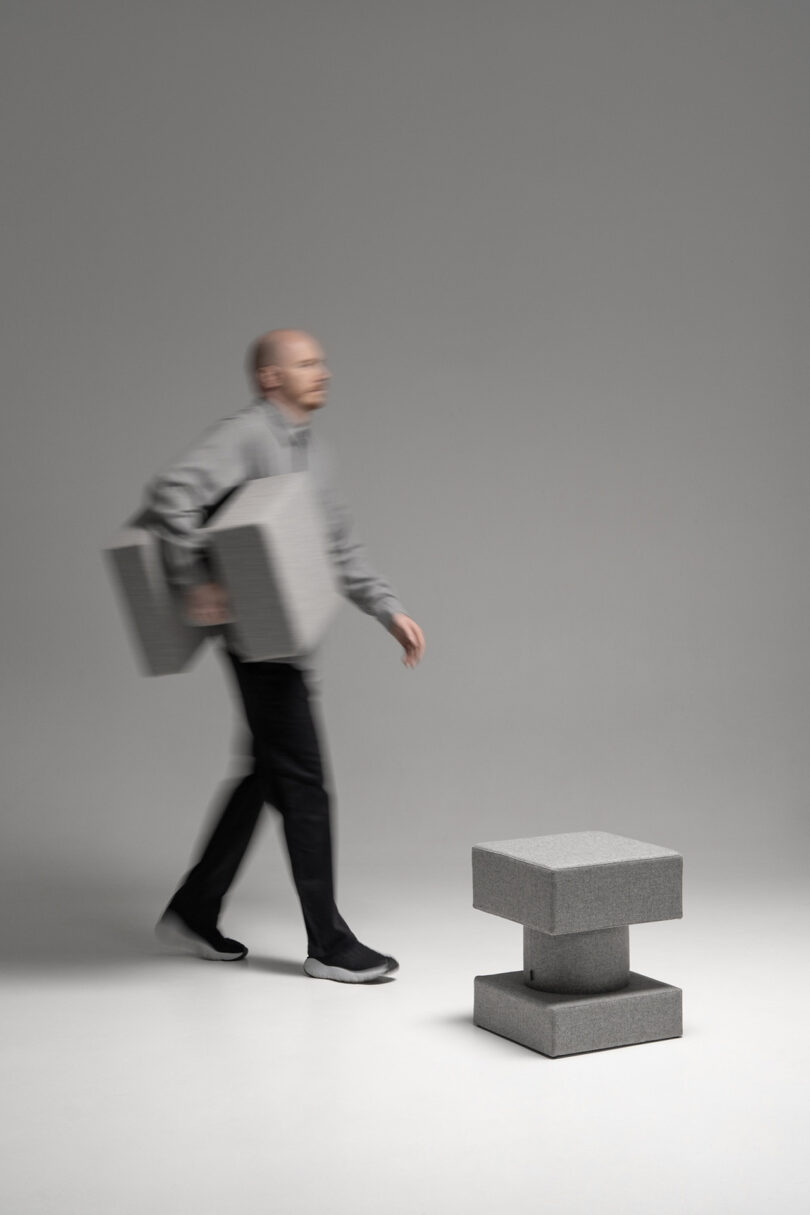 A person carries two large gray foam blocks past a matching stacked block structure on a plain gray background.