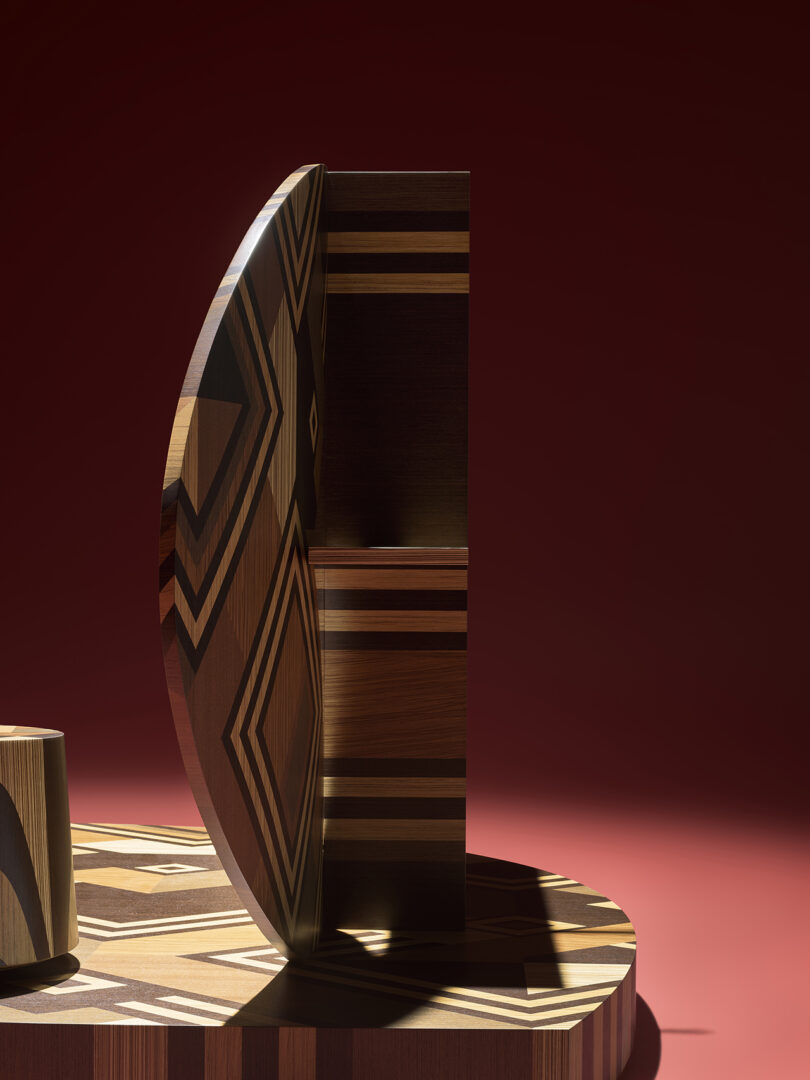 A geometric wooden sculpture featuring a semi-circular panel with patterned inlays, set against a red background with a matching striped base.