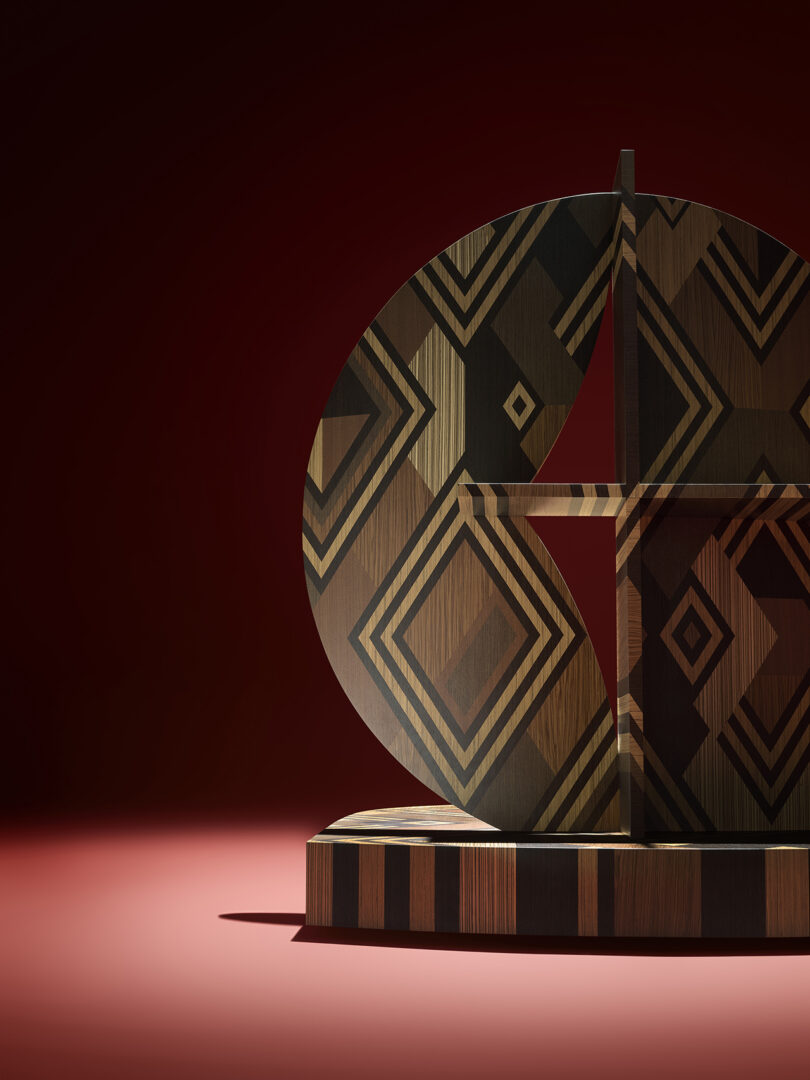 A geometric wooden sculpture with diamond and stripe patterns stands on a round base, set against a red background with dramatic lighting.