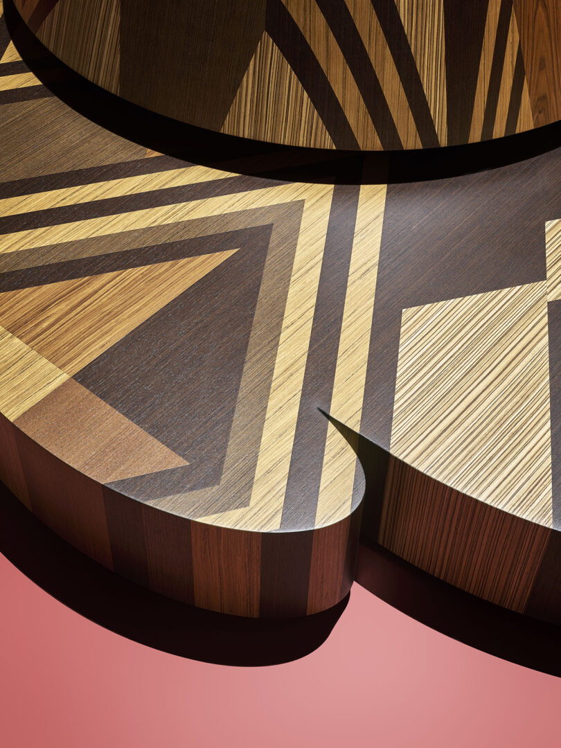 Geometric wooden shapes with inlaid patterns in various wood tones rest on a solid pink background.