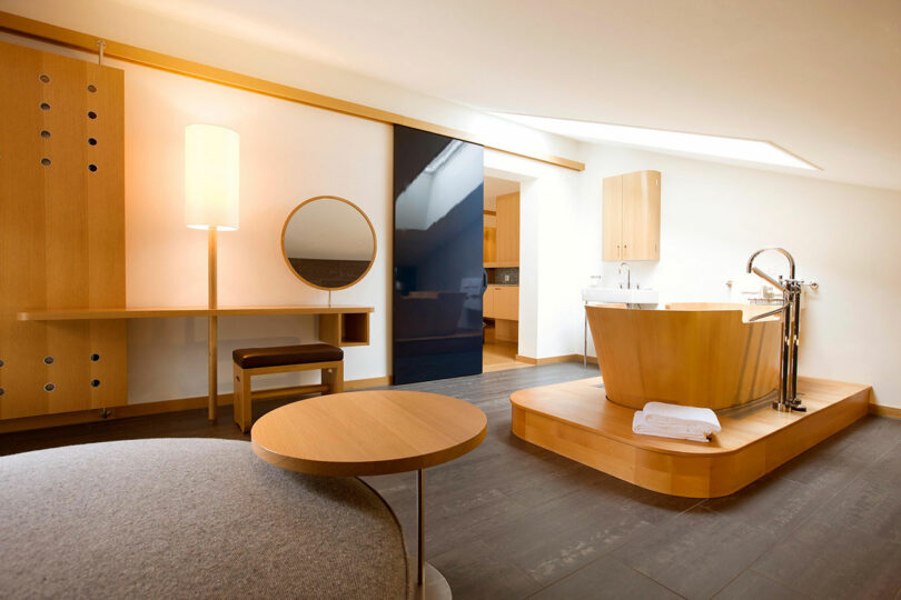 El baño moderno cuenta con una bañera de madera sobre una plataforma, un espejo redondo sobre el banco, una lámpara de pie y muebles minimalistas de madera.