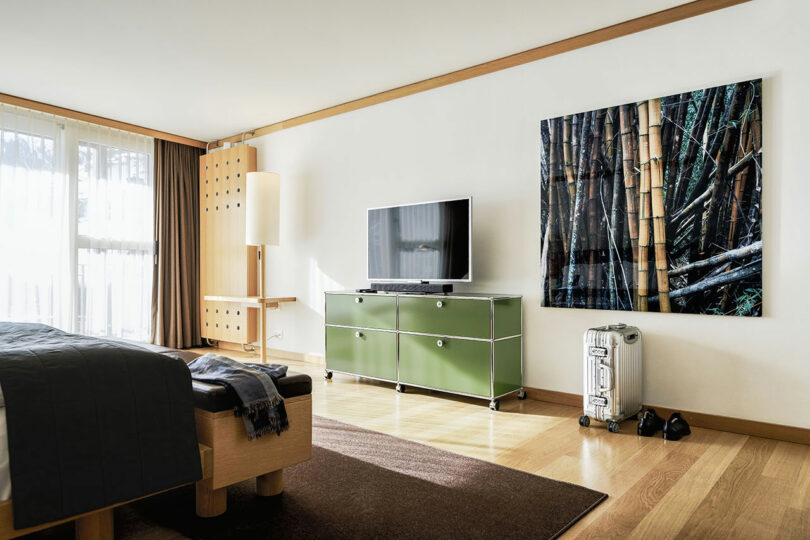 Moderna habitación de hotel con cama, TV en una cómoda verde, grandes obras de arte de bambú en las paredes, maleta, zapatos y ventanales con cortinas del suelo al techo.