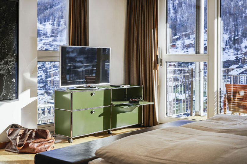 El moderno dormitorio cuenta con un mueble de televisión verde, un televisor de pantalla plana, una gran ventana con cortinas marrones y vistas a los edificios cubiertos de nieve y a las montañas del exterior.