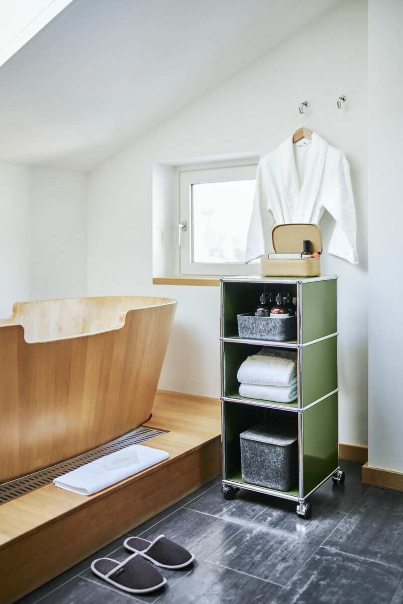 El moderno cuarto de baño cuenta con una bañera de madera, un carrito verde con toallas y artículos de tocador, albornoces blancos colgados de ganchos, una ventana y zapatillas sobre el suelo de baldosas oscuras.