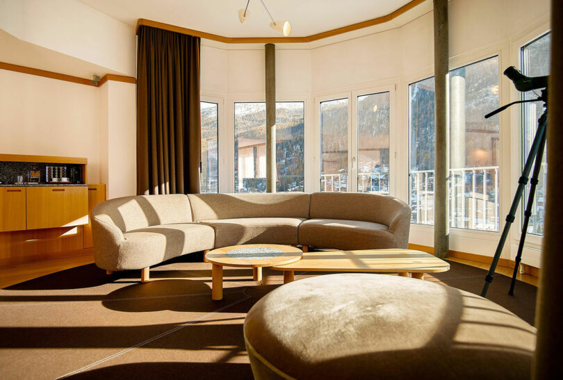 Salón moderno con sofá curvo beige, taburete redondo, mesa de centro de madera, ventana grande y telescopio junto a la ventana.