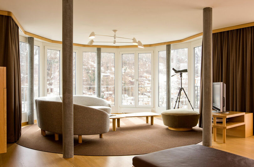 Salón moderno con sofá curvo, taburete redondo, TV, telescopio y ventanales con vistas al paisaje nevado. La combinación de colores marrón y beige se combina con suelos de madera.