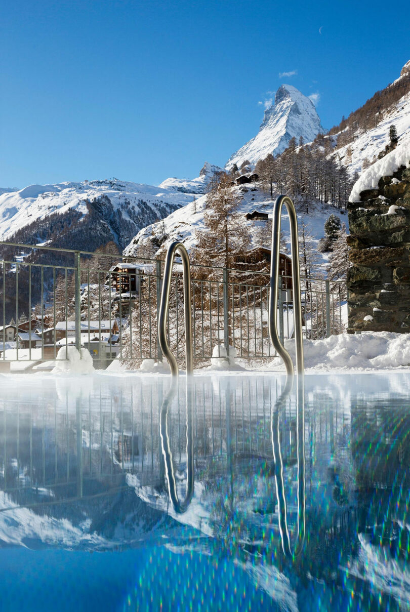 Piscina al aire libre con escalera de metal en un paisaje nevado bajo un cielo azul claro con Matterhorn visible en el fondo.