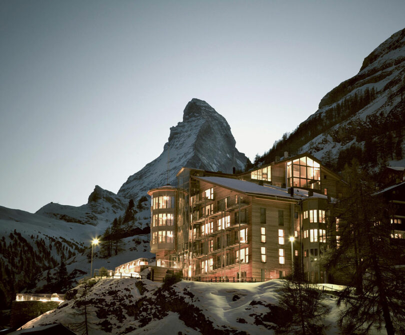 Un moderno edificio de hotel de madera iluminado al atardecer con el telón de fondo de montañas cubiertas de nieve y el Matterhorn al fondo.