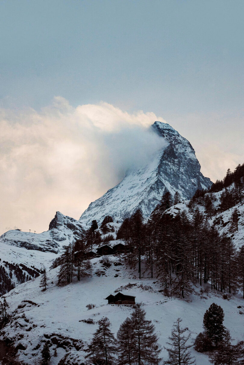 Un pico de montaña cubierto de nieve, parcialmente oscurecido por las nubes, con una cabaña y escasos árboles en primer plano.