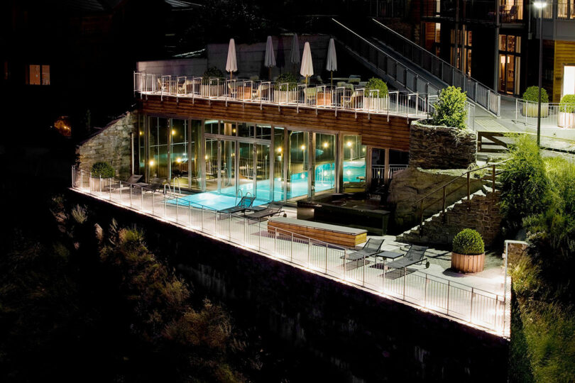 La moderna piscina exterior y la terraza nocturna están iluminadas con luces y equipadas con paredes de cristal, tumbonas y mesas con sombrillas.