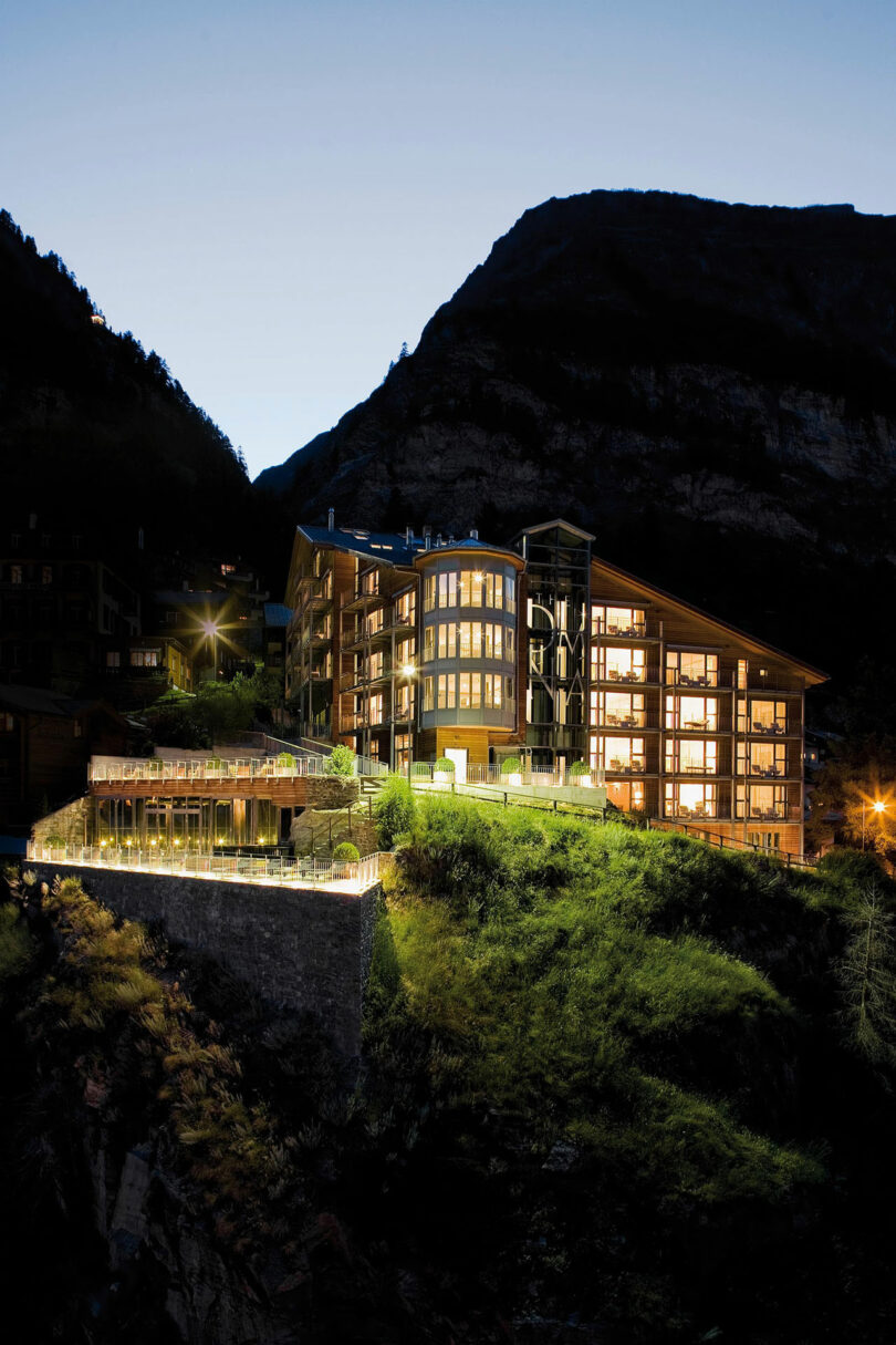 Este moderno hotel de varios pisos está ubicado en una ladera con las montañas al fondo y sus grandes ventanales se iluminan al anochecer.