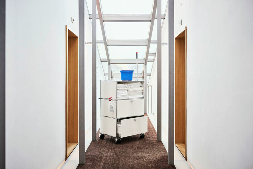 Un camión de limpieza cargado con toallas dobladas y un bote de basura azul está estacionado en un pasillo bien iluminado con paredes blancas y alfombra marrón.