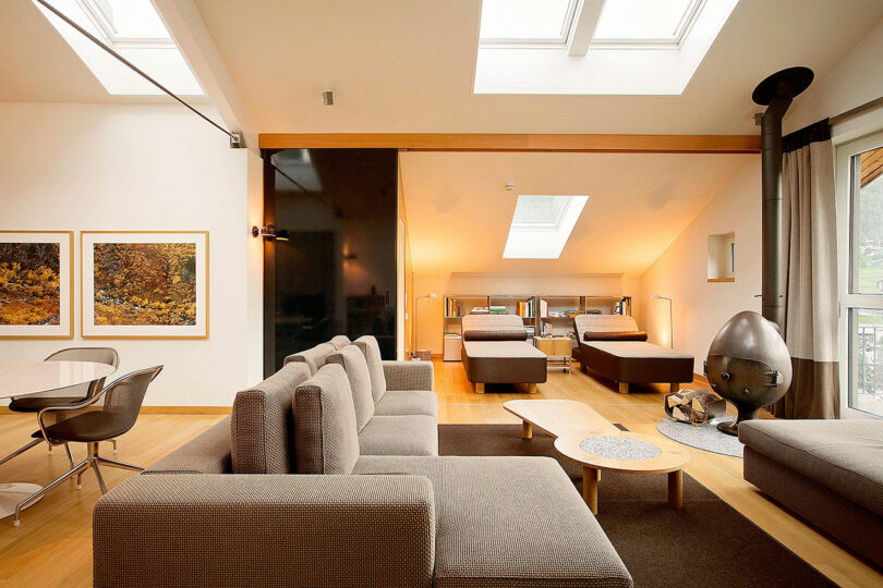 Moderna sala de estar de planta abierta con sofá seccional gris, mesa de comedor con sillas, dos camas, claraboya, estufa de leña y fotografías de paisajes enmarcadas en la pared.