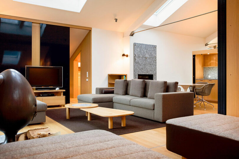 Sala de estar moderna con sofá gris, mesa de centro de madera, TV de pantalla plana y vista abierta al comedor y la cocina. Iluminación natural procedente de suelos de madera clara y claraboyas.