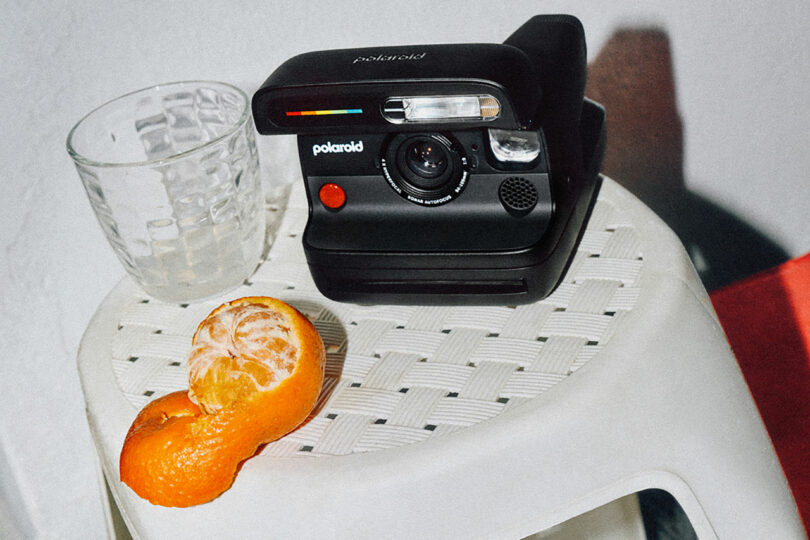 Una cámara instantánea Polaroid negra, un trozo de vidrio transparente y una naranja parcialmente pelada descansan sobre un taburete tejido blanco.