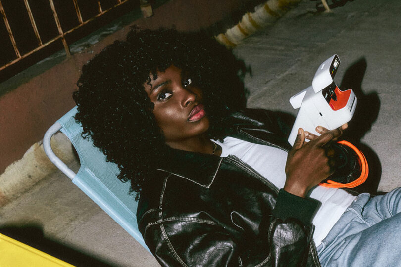 En un ambiente al aire libre, un hombre con cabello rizado y una chaqueta de cuero negra se sienta en una silla, sostiene una cámara de película instantánea y mira a la cámara.