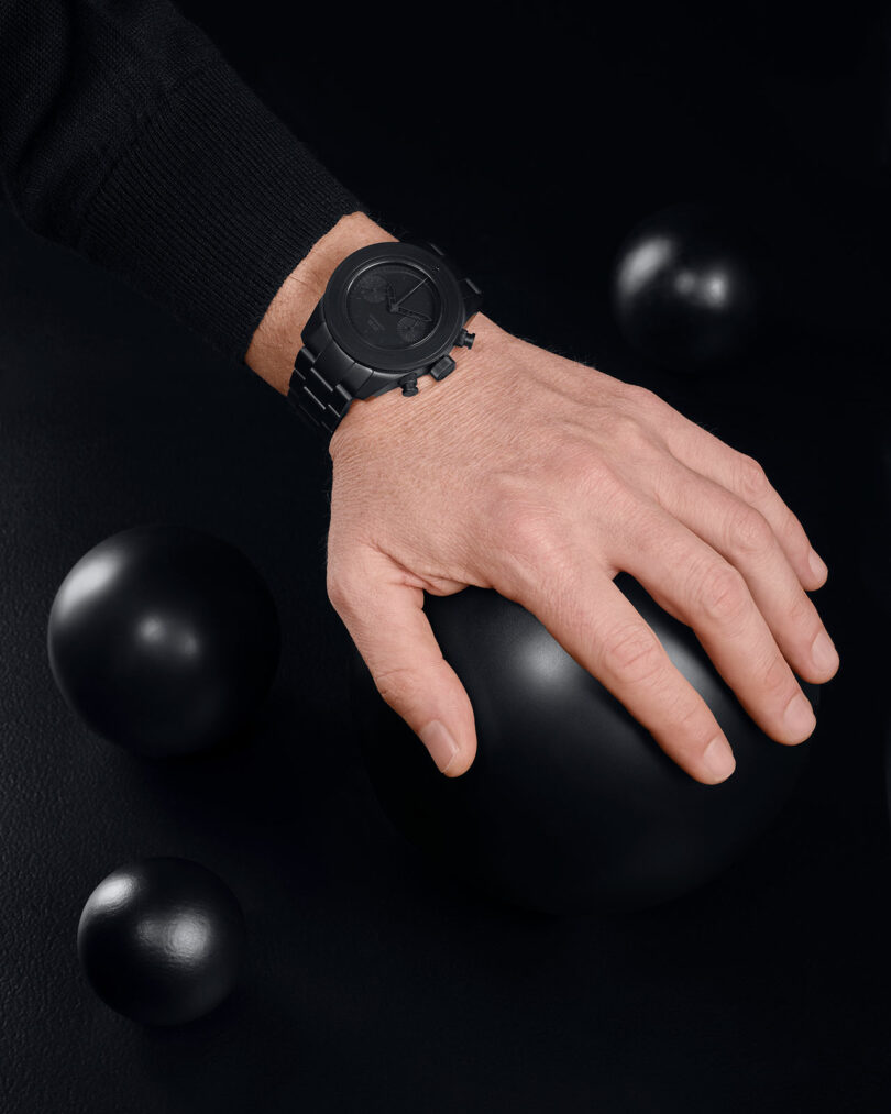 A hand wearing a black wristwatch rests on a large black sphere, surrounded by smaller black spheres on a dark surface.