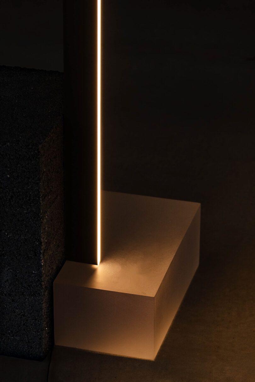 En el ambiente oscuro, una franja vertical de luz cálida se encuentra entre bloques de textura oscura y una base rectangular suave de color beige.