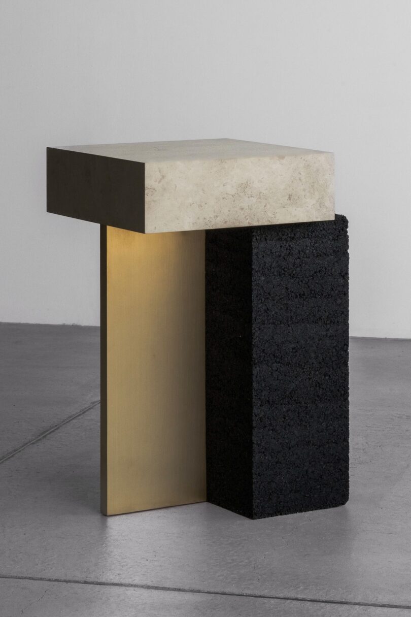 Una mesa auxiliar moderna con tapa de piedra beige, paneles verticales de latón y bloques negros texturizados como soporte contrasta con las paredes grises lisas y el piso de concreto.