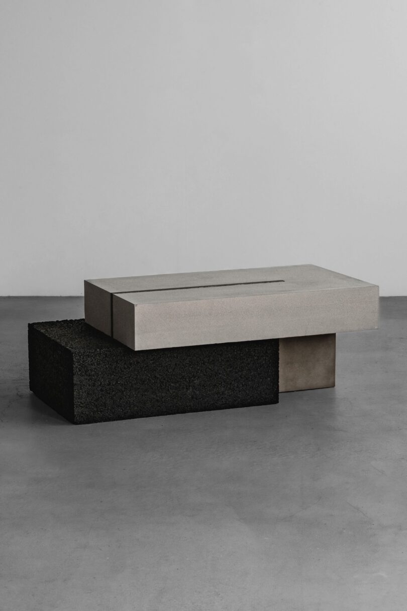 Una mesa de centro minimalista con dos secciones de bloques rectangulares, una gris claro y otra negra, está colocada sobre un piso de concreto liso contra una pared lisa.