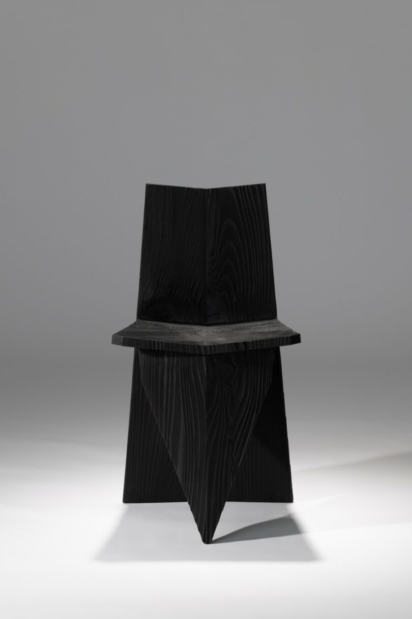 Una silla de madera negra y angular con formas geométricas y líneas nítidas, centrada en un fondo gris sólido.