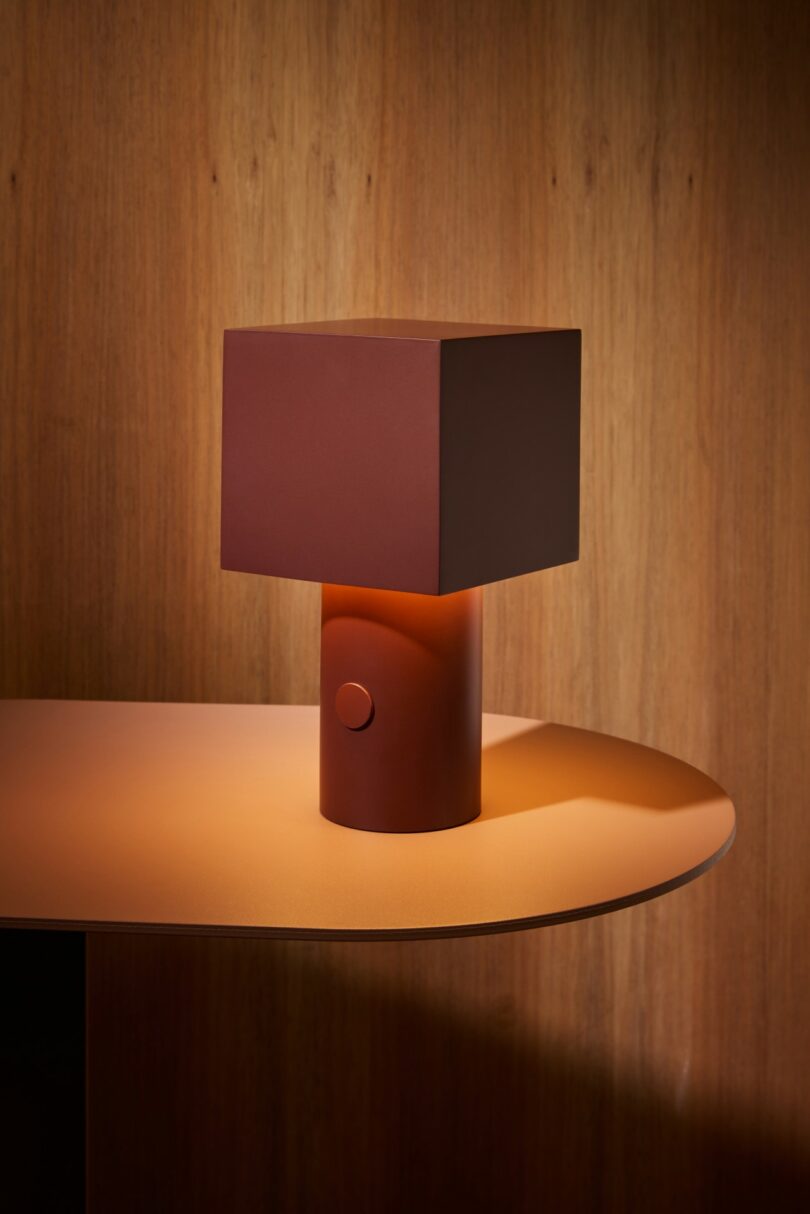 A modern table lamp with a square, brown shade and cylindrical base sits on a curved, light brown surface against a wooden background