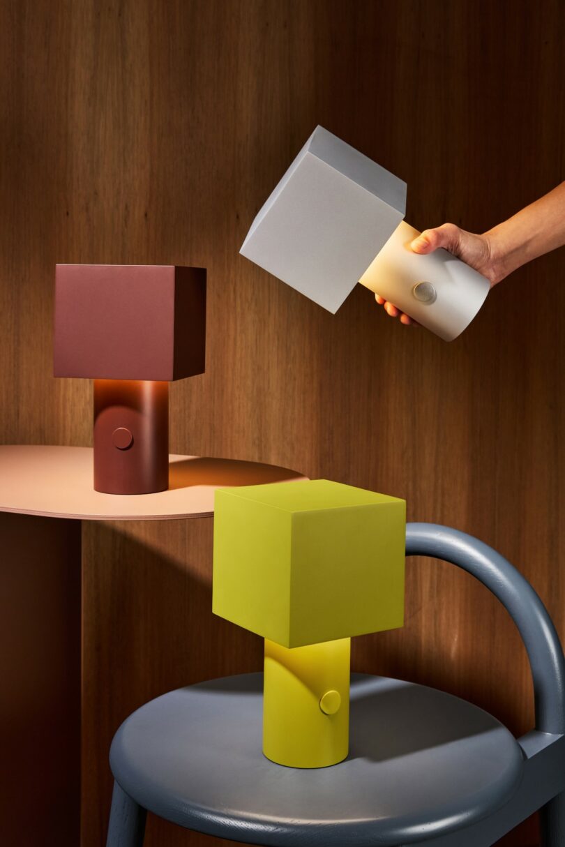 Three geometric, cube-shaped table lamps in brown, yellow, and white are displayed on furniture against a wooden background; a hand lifts the white lamp