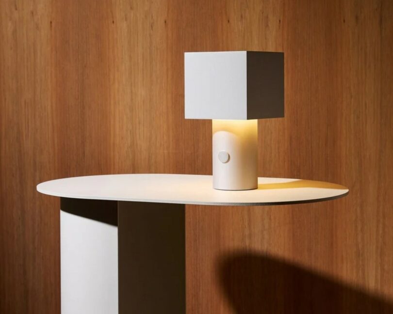 A square white table lamp with a cylindrical base sits on a minimalist oval table against a wooden wall background