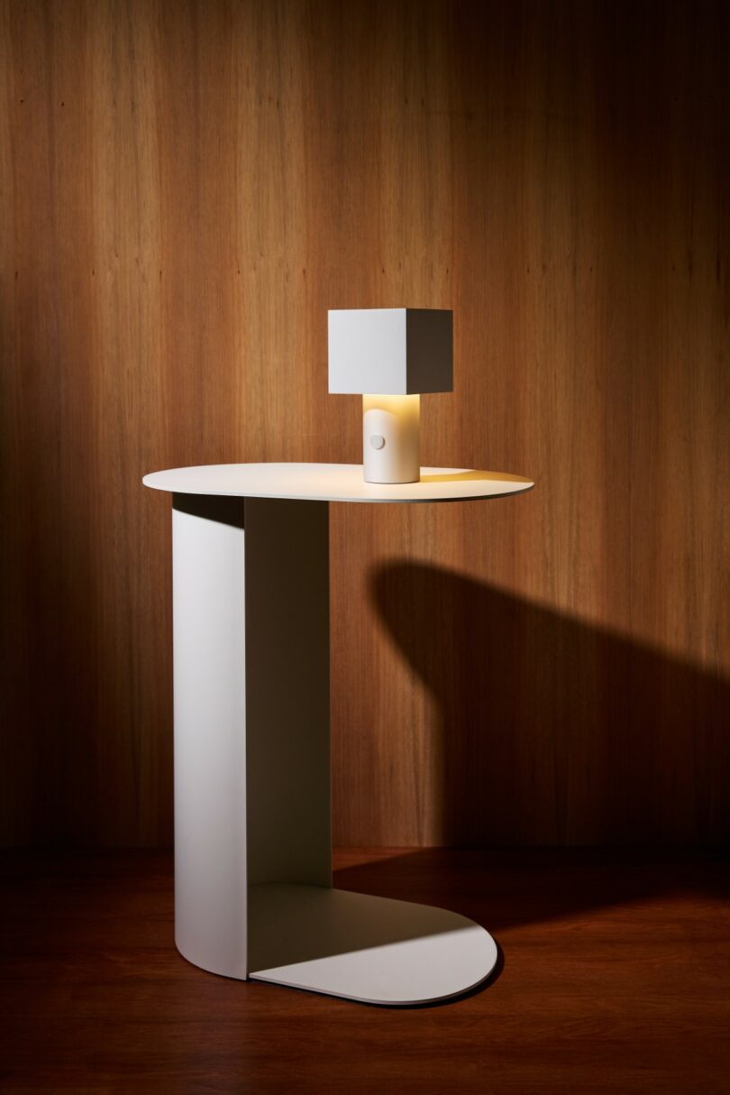 A minimalist white side table with a curved base holds a small, modern lamp with a square shade, set against a wood-paneled wall