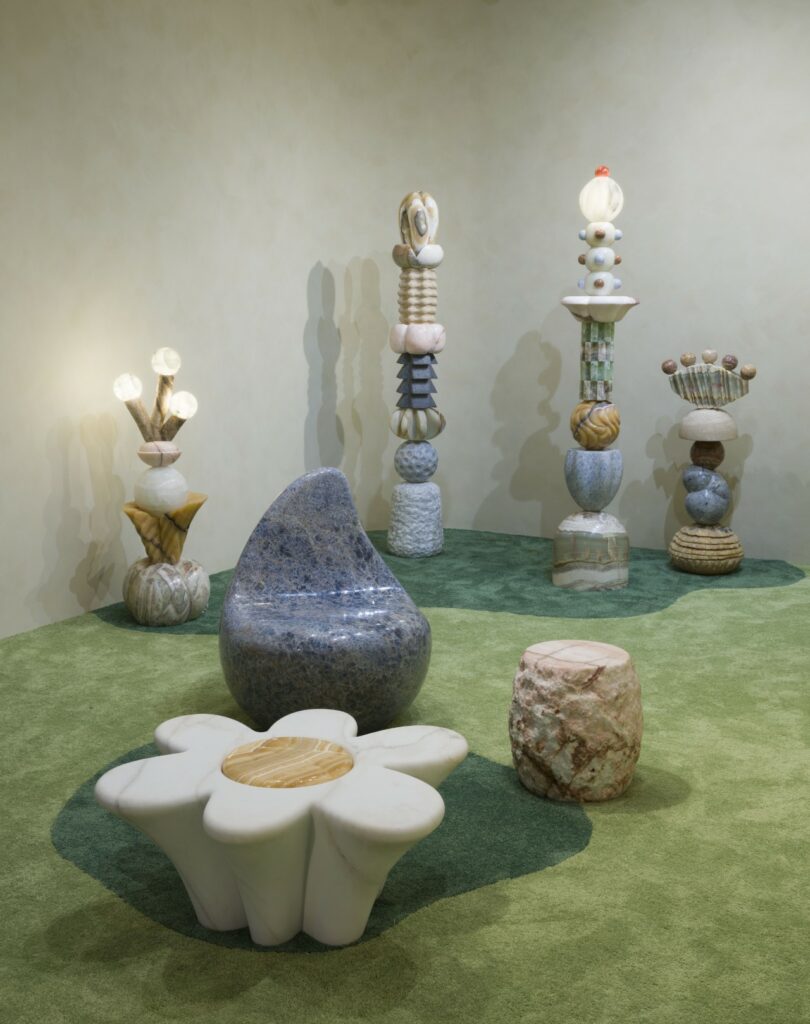 La sala alfombrada de verde presenta esculturas abstractas y muebles de piedra de Sten Studio, incluida una mesa con forma de flor, una silla azul y varias estructuras en forma de tótem con elementos esféricos.