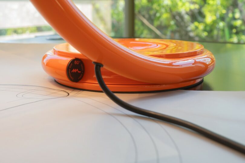 Primer plano de la base de una lámpara de mesa naranja sobre una mesa con cable de alimentación visible y dibujos técnicos; plantas verdes en el fondo borroso