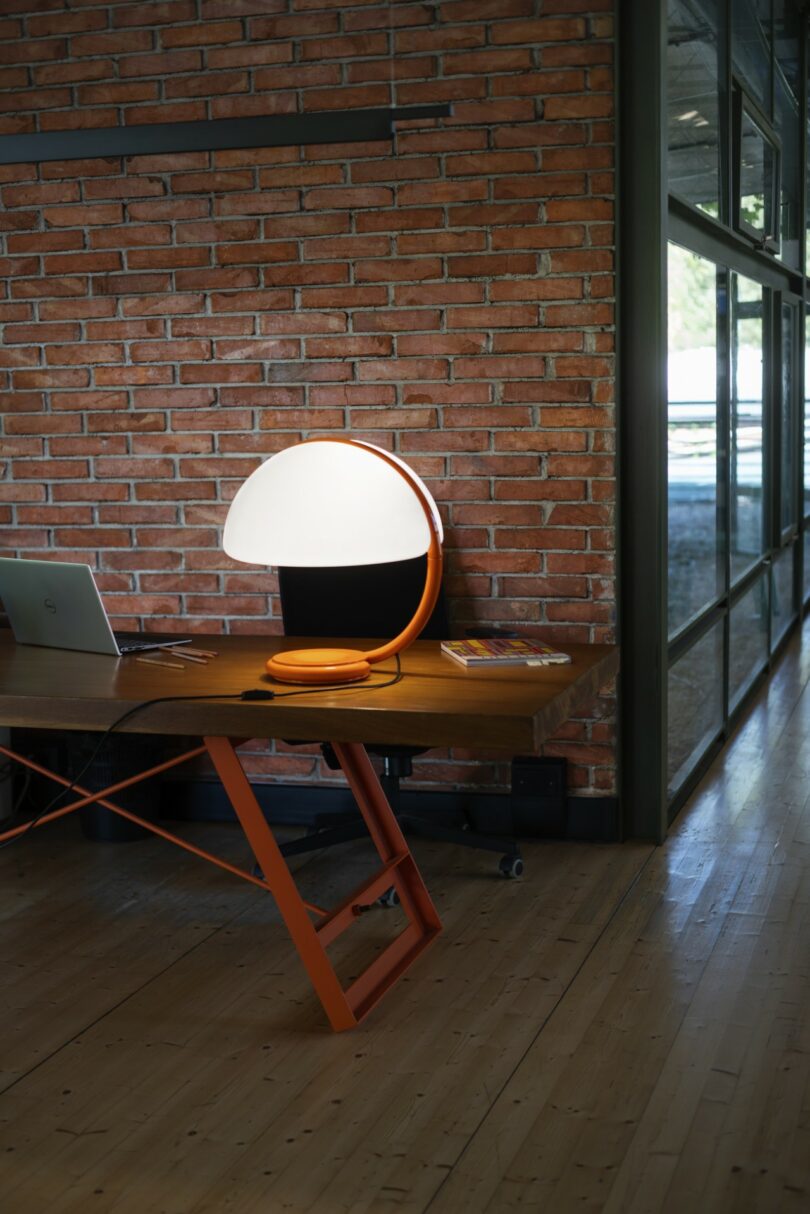 Una moderna lámpara de mesa naranja ilumina una mesa de madera con un portátil cerrado y libros contra una pared de ladrillo rojo y una gran ventana de cristal