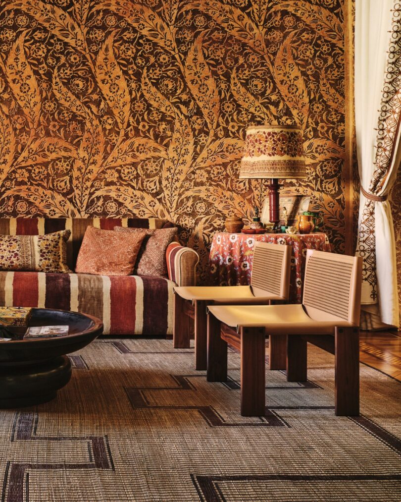 Acogedor salón con paredes de tapices estampados, sofá a rayas, lámpara decorativa, mesa redonda, dos sillas modernas y alfombra tejida. Los cálidos tonos tierra dominan el espacio.