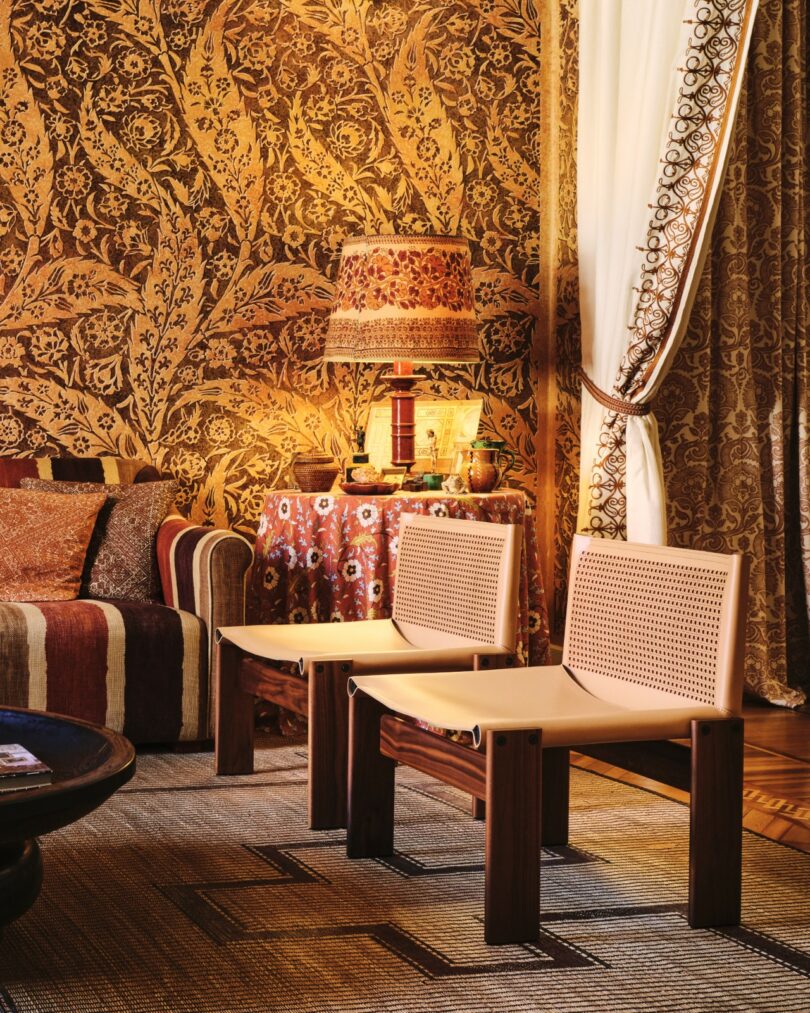 Salón con papel pintado marrón estampado, sofá con cojines a rayas, dos sillas de madera, una lámpara sobre una mesa redonda cubierta con tela estampada y cortinas con ribetes bordados