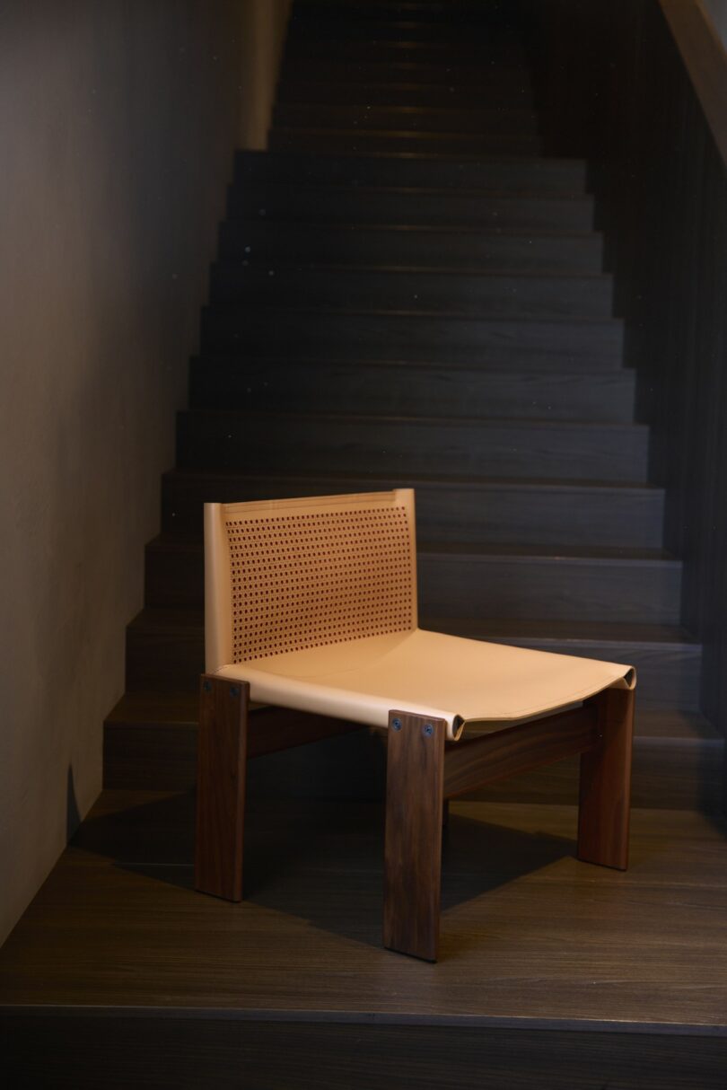 Una moderna silla de madera con respaldo y asiento de cuero perforado se coloca en el suelo de madera oscura frente a las escaleras.