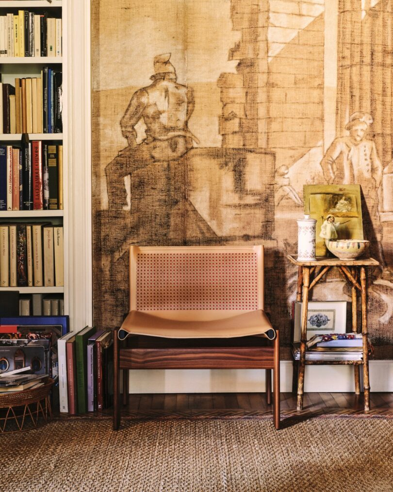 Una silla de madera con asiento de mimbre se encuentra junto a una pequeña mesa auxiliar con libros, cerámica y objetos de arte, frente a un mural y una estantería llena de libros.