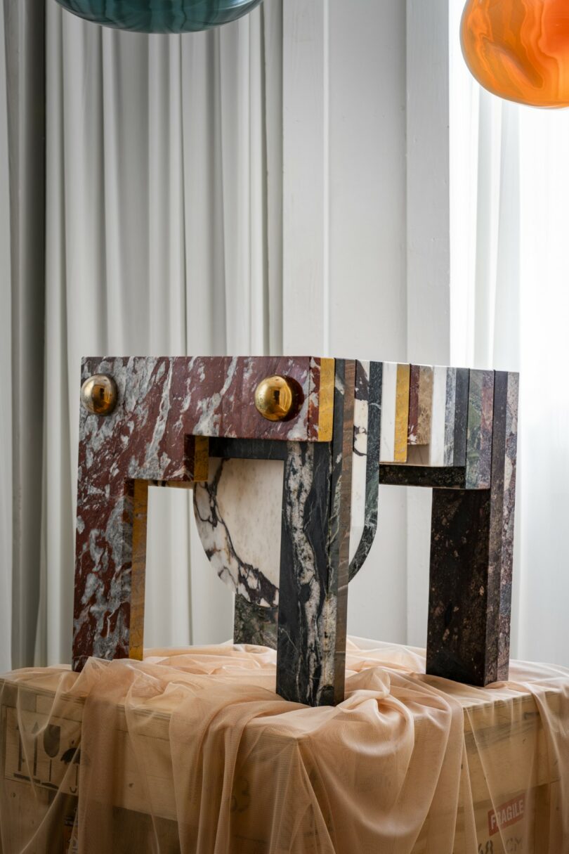 Una colorida mesa de mármol con formas geométricas y dos pomos redondos dorados se coloca en una habitación cubierta de cajas con paredes de paneles blancos y lámparas de cristal colgantes.