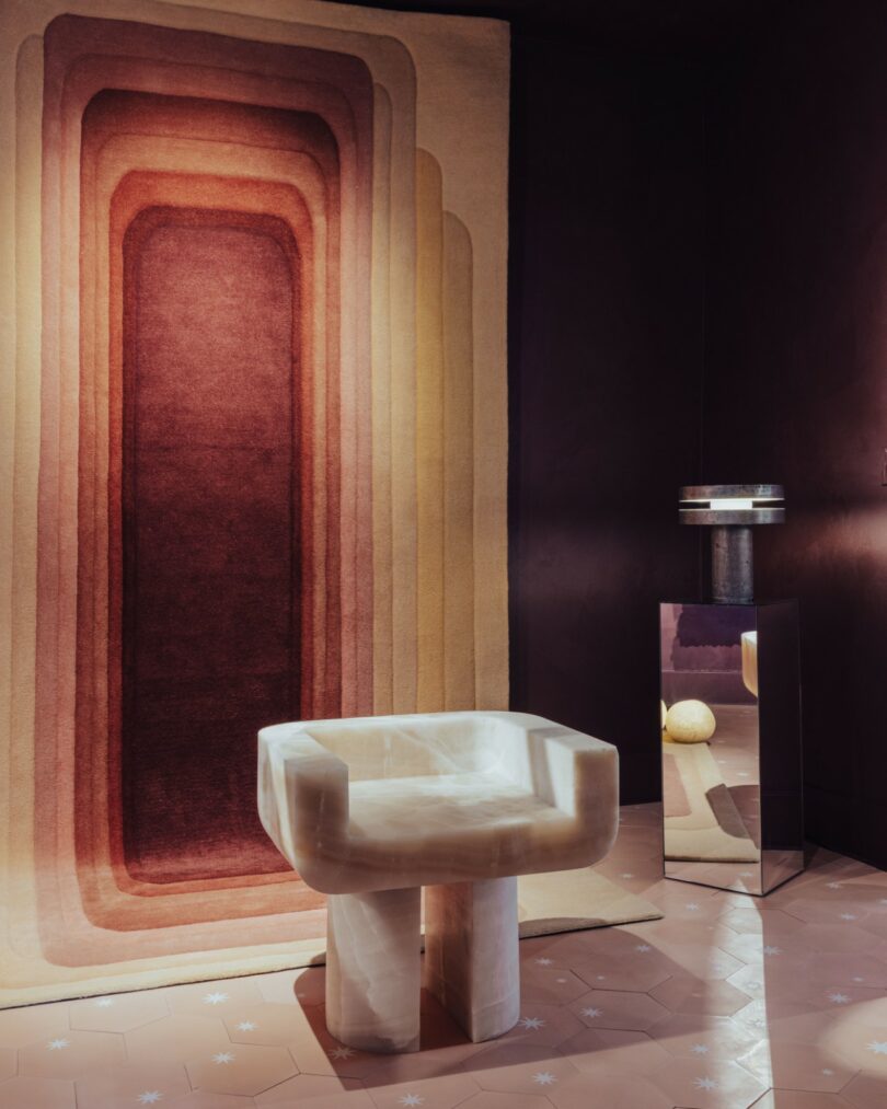 La habitación contemporánea cuenta con sillas geométricas de color beige, una alfombra alta degradada en tonos marrones y rojos, una mesa baja con espejos y un piso de baldosas hexagonales de color arcilla.