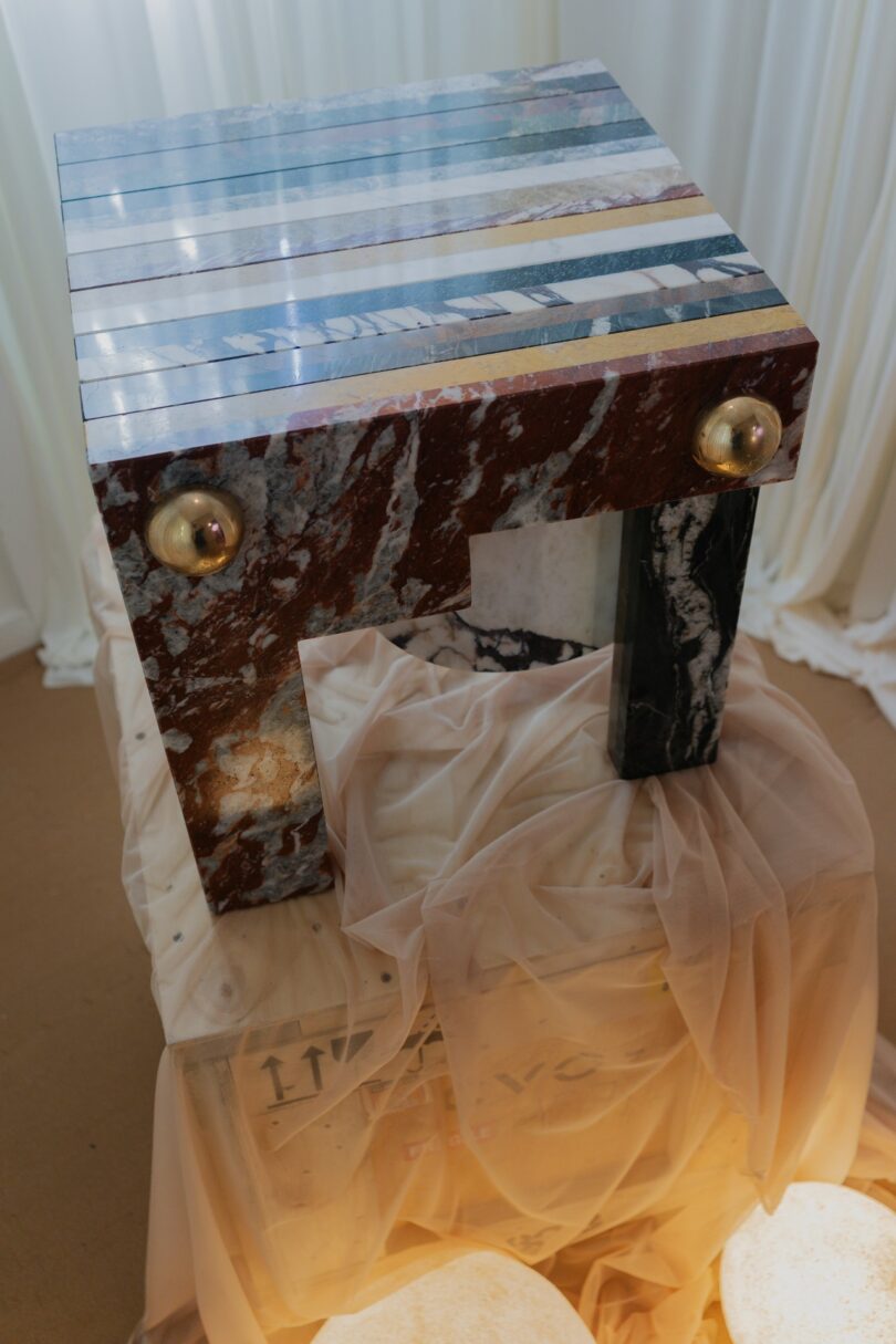 Una mesa geométrica con una superficie decorada con piedras de rayas de colores y esferas doradas descansa sobre una caja de madera cubierta con tela translúcida y rodeada de luces suaves.