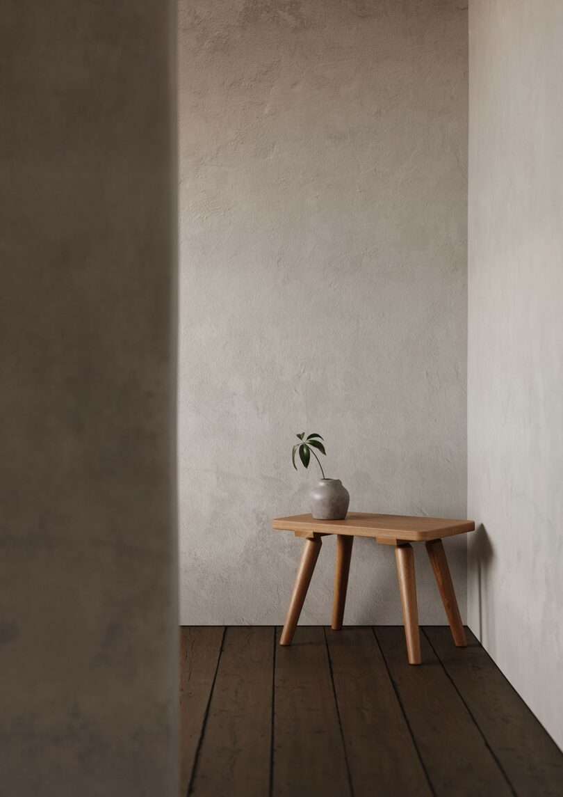 Una pequeña mesa de madera de cuatro patas descansa contra una pared beige texturizada con una maceta de hojas verdes; el suelo de madera oscura es claramente visible