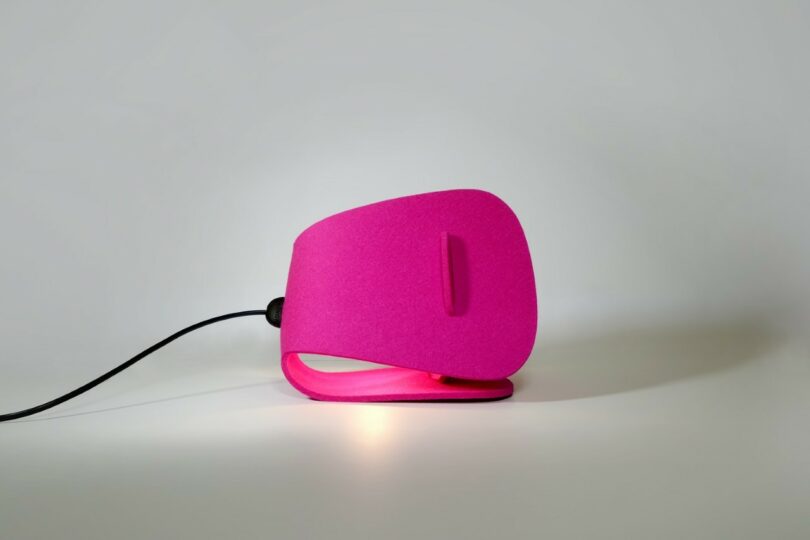 Una lámpara de mesa moderna y angular de color rosa que emite un brillo suave con un diseño geométrico y un cable de alimentación visible sobre un fondo liso.