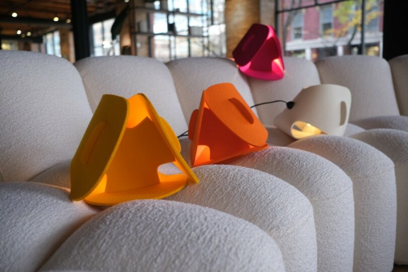 Cuatro lámparas geométricas amarillas, naranjas, rosas y blancas colocadas sobre un sofá de textura blanca en una moderna habitación iluminada por el sol