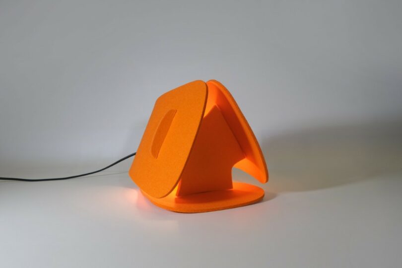 Lámpara de mesa moderna y angular de color naranja con brillo suave y diseño geométrico con cable de alimentación visible sobre un fondo liso.