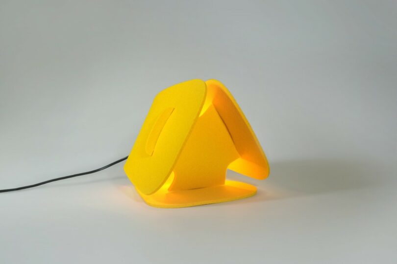 Lámpara de mesa moderna y angular de color amarillo con brillo suave, diseño geométrico, cable de alimentación visible y fondo liso.