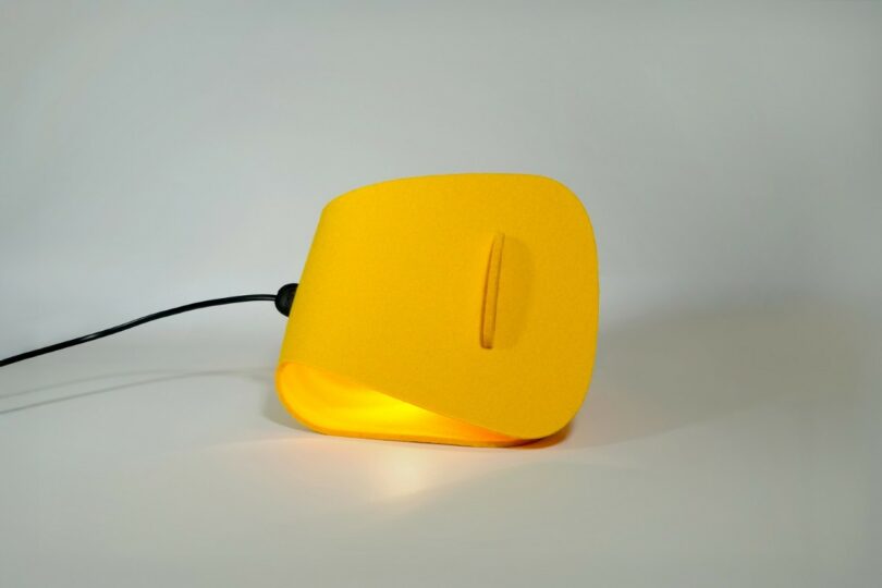 Lámpara de mesa moderna y angular de color amarillo con brillo suave, diseño geométrico, cable de alimentación visible y fondo liso.