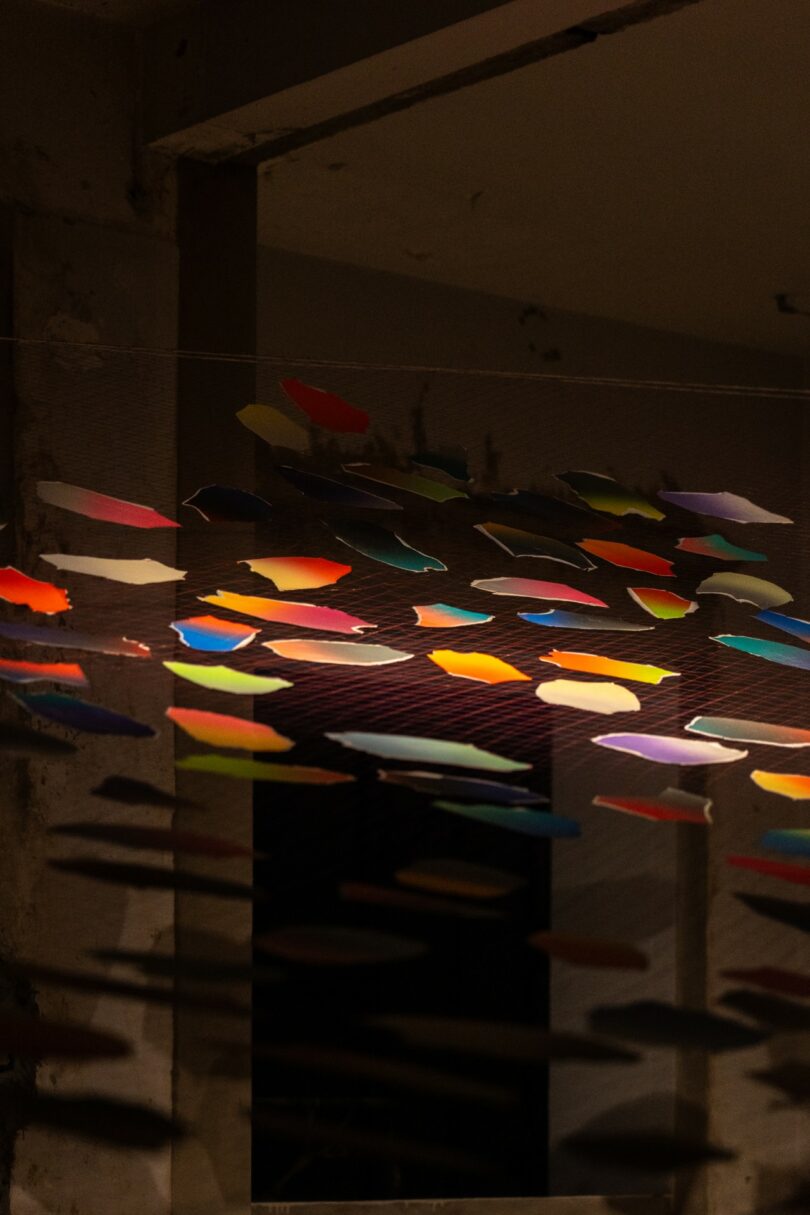 Coloridas formas de papel que cuelgan de cuerdas en el interior, proyectando sombras en las paredes y el techo en una habitación con poca luz y vigas de hormigón expuestas