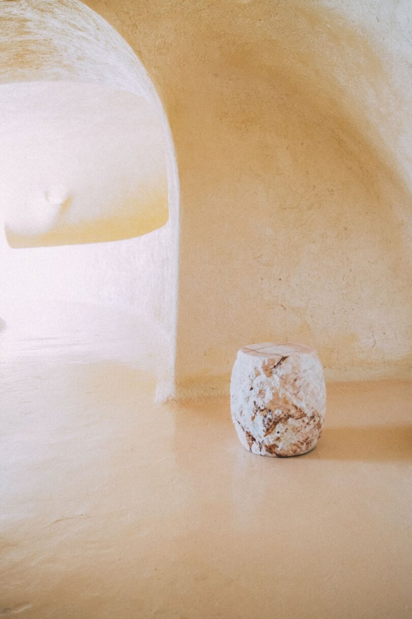 Un banco de piedra redondo y texturizado se asienta sobre un piso liso de color melocotón en una habitación curva y minimalista con paredes enlucidas y una abertura arqueada.