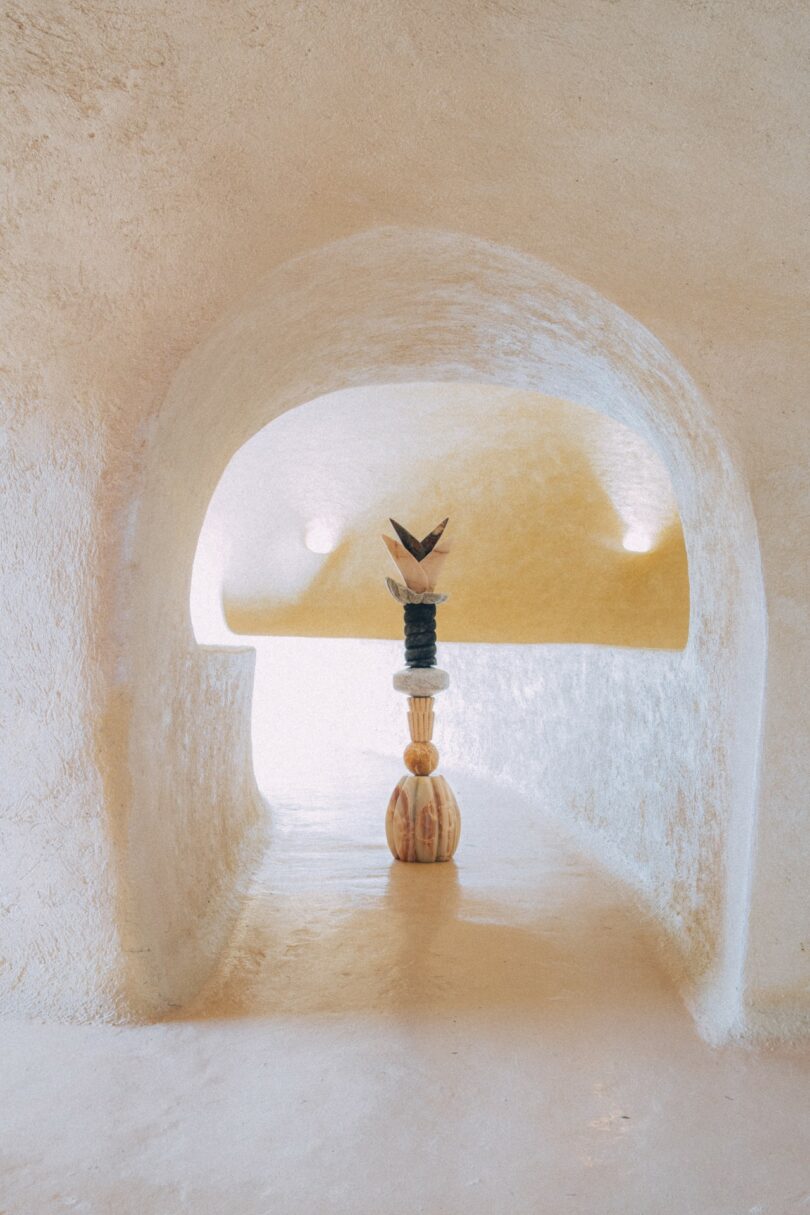 Una obra de arte escultórica con formas geométricas apiladas se encuentra en el centro de un elegante corredor arqueado de color beige con una luz suave.