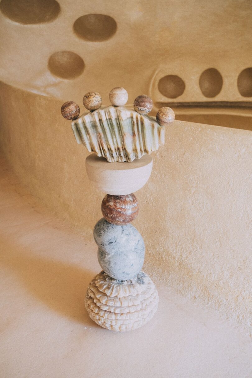 Piedras y cerámicas redondas y texturizadas se apilan sobre suelos de color beige y se disponen escultóricamente sobre paredes curvas de color arena con hendiduras circulares.