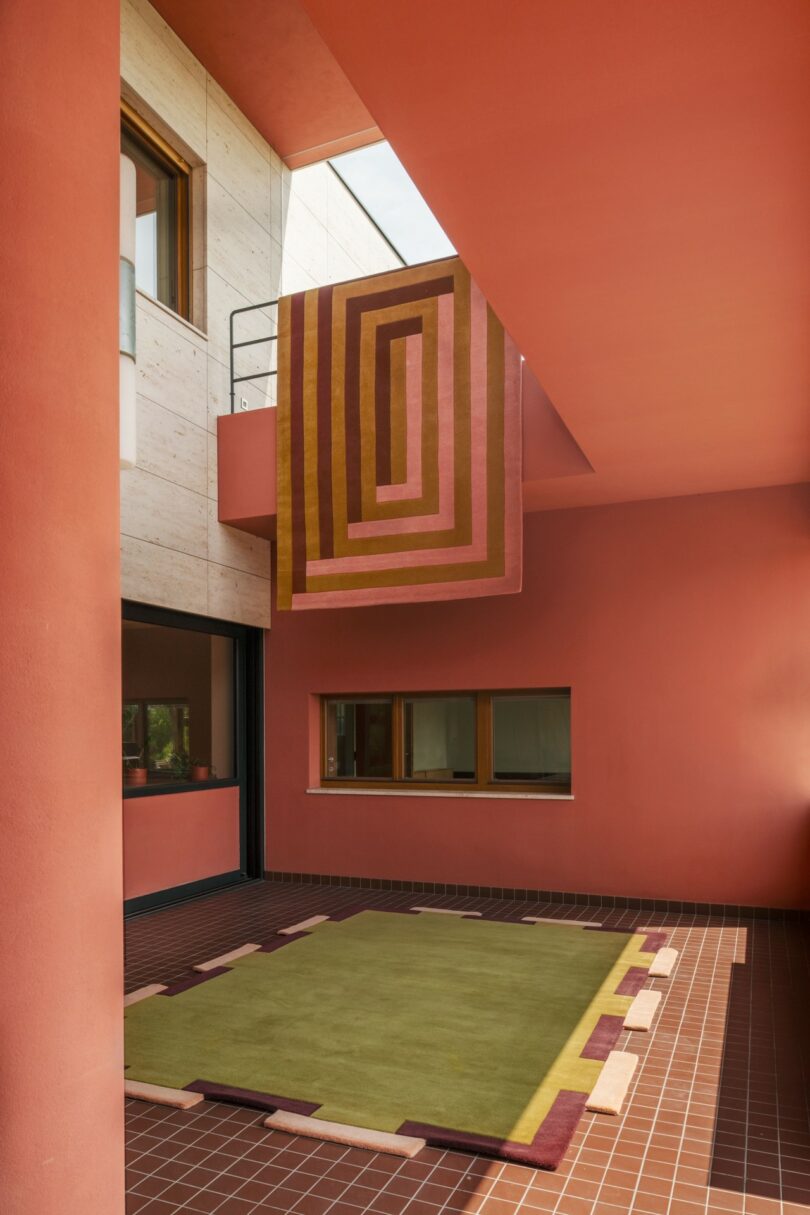 Interior de estilo moderno con paredes de color salmón, suelos de baldosas, una gran alfombra verde y textiles colgantes con motivos geométricos en colores marrón, rosa y oliva.